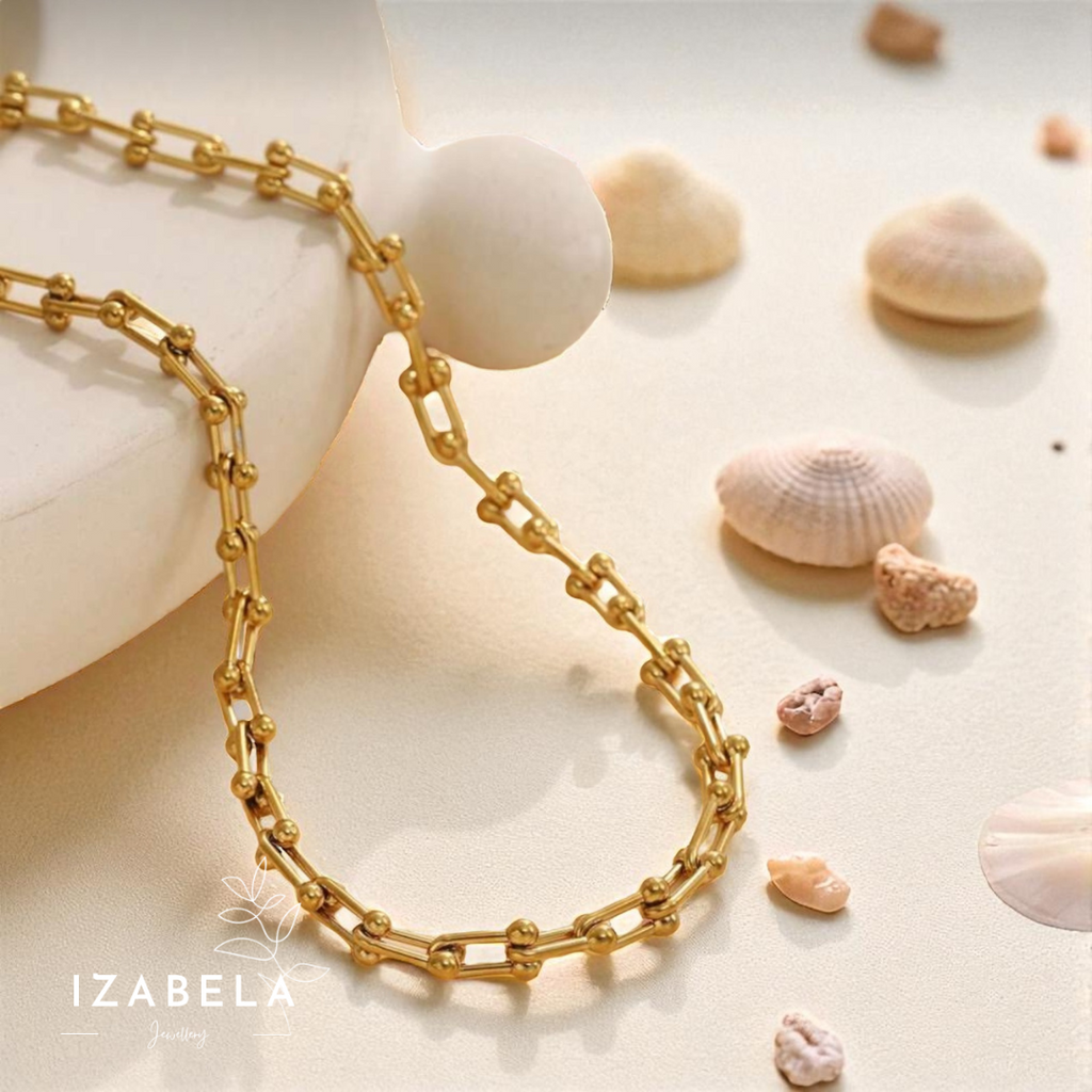 Gold chain necklace on a sandy surface with seashells, featuring the brand 'IZABELA'.