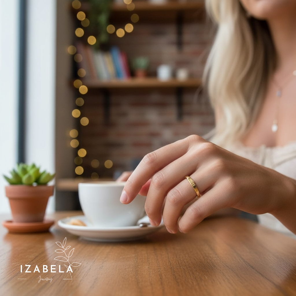Person wearing a gold ring on a wooden table with a cup and saucer, blurred background with lights and books.